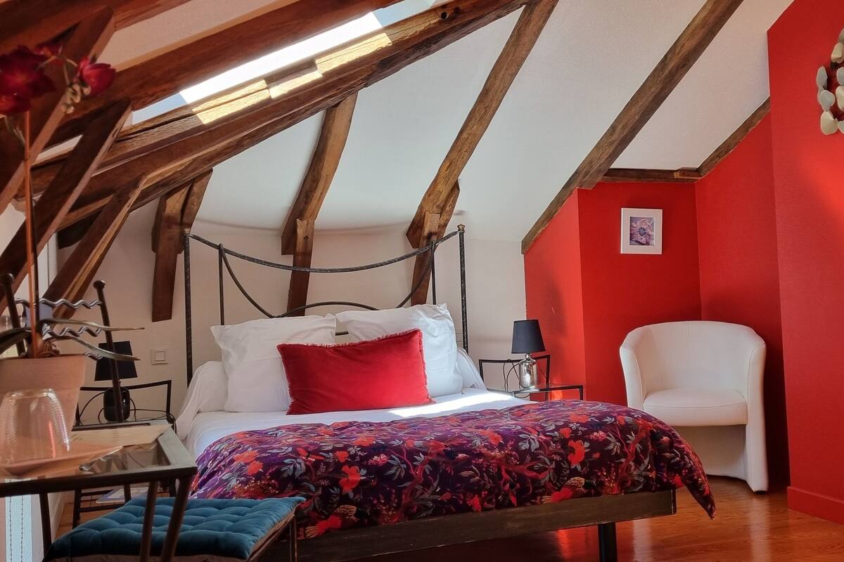 Ein Schlafzimmer mit schrägen Wänden, einem Bett mit floralem Überwurf, einem weißen Stuhl und einer roten Wand.