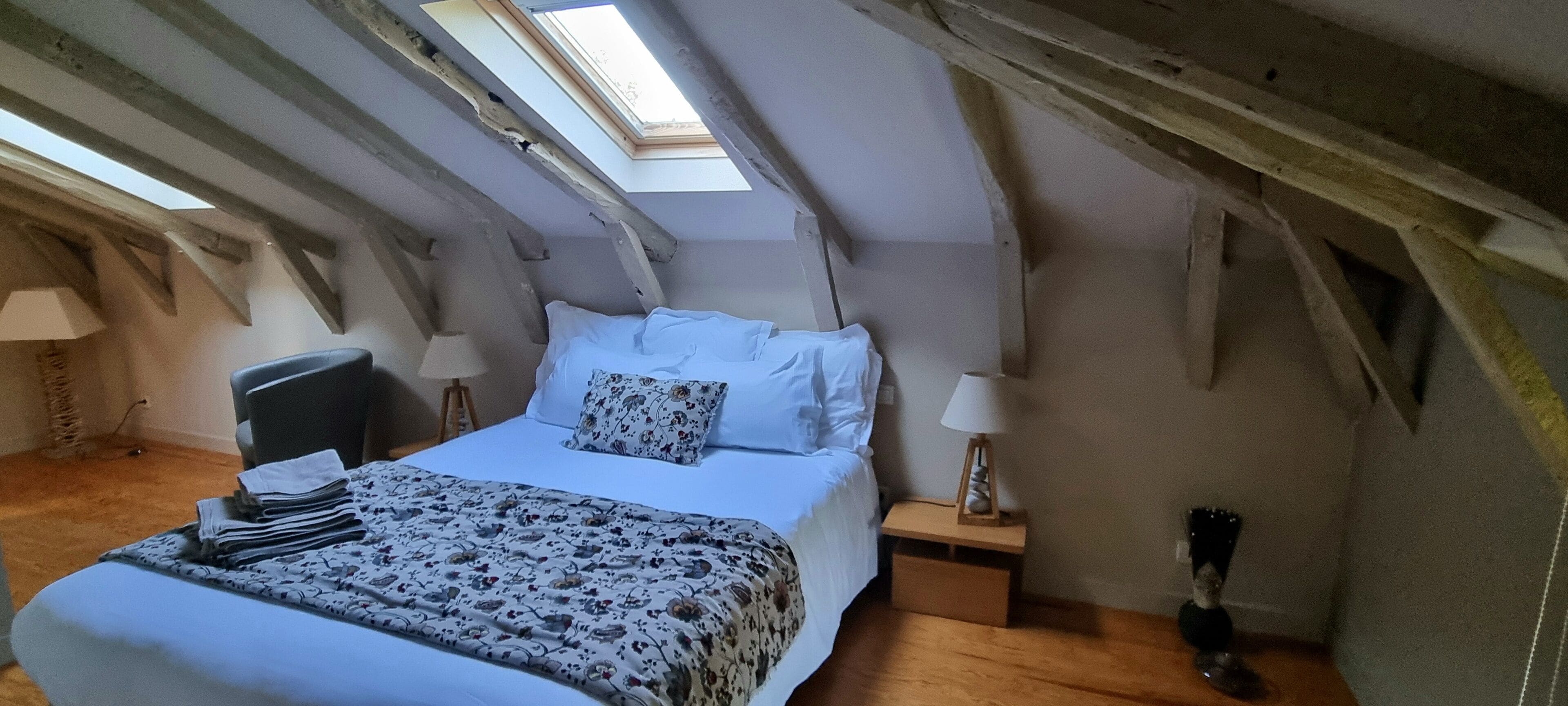 Une chambre avec un puits de lumière, un lit avec une literie à motifs floraux, une chaise, des tables de chevet avec des lampes et un plancher en bois.
