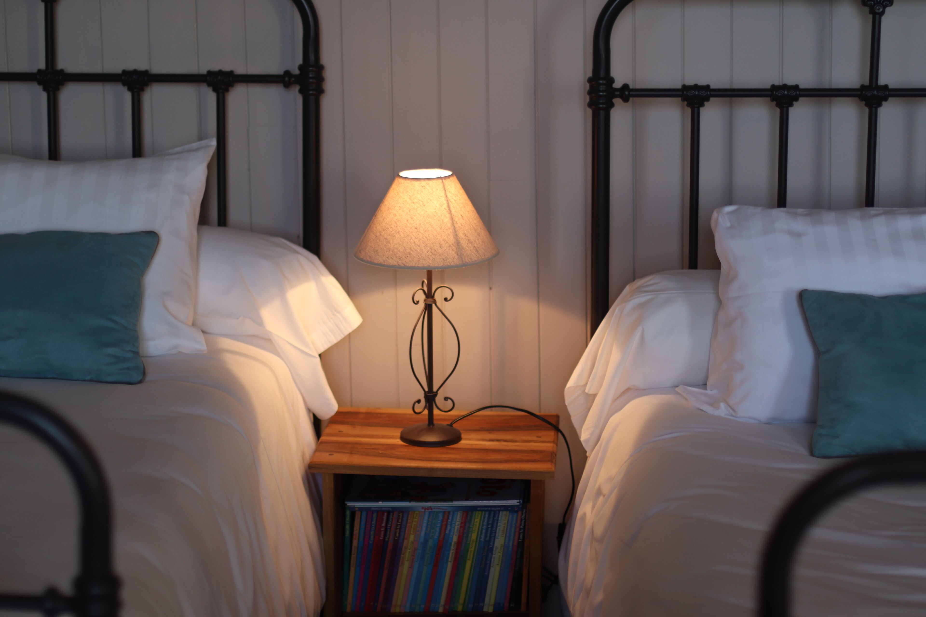 Une chambre avec deux lits, chacun agrémenté de draps blancs et d’oreillers turquoise. Une table de chevet avec une lampe et une bibliothèque se trouve entre les deux lits.