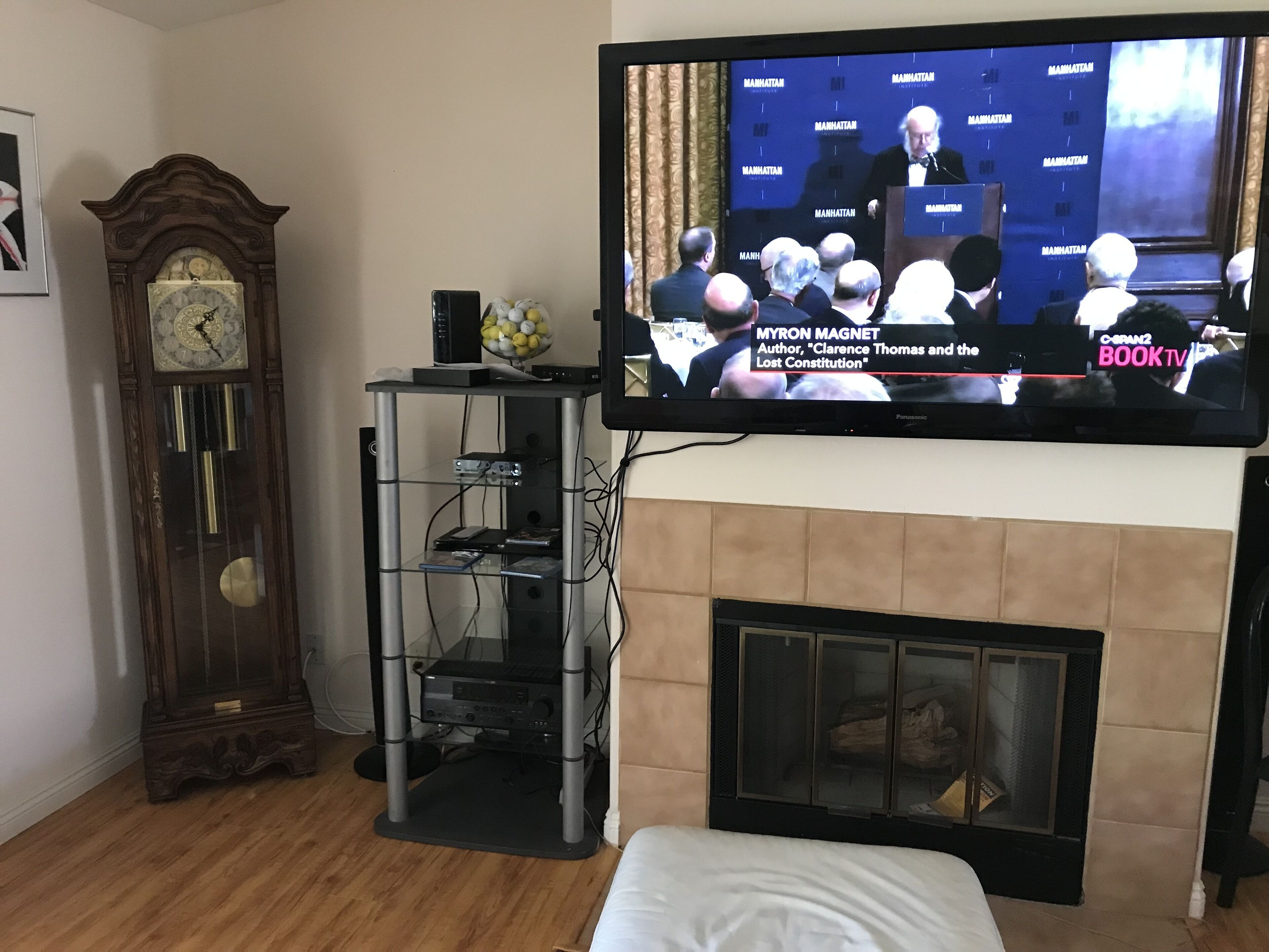 A television mounted on a wall displaying a news broadcast, a wooden grandfather clock, and a fireplace with a modern entertainment unit below.