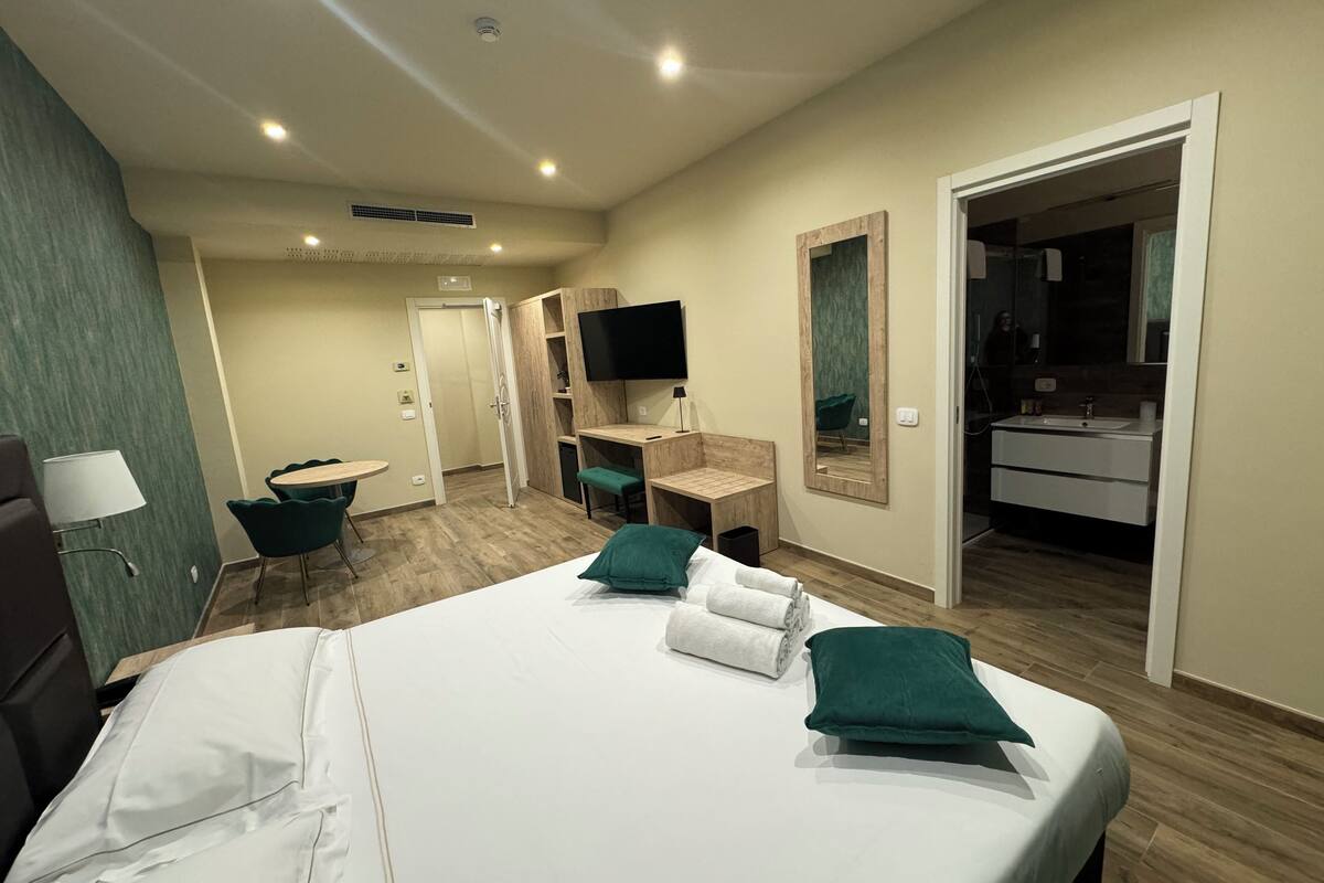 A hotel room with a bed, a desk, a chair, a TV, a mirror, and a bathroom.