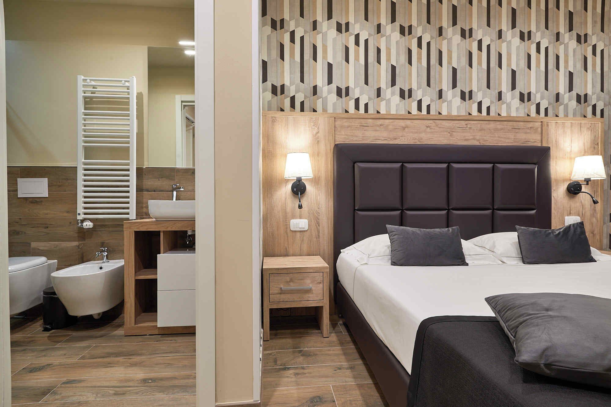 Habitación de hotel con cabecera de madera, una cama grande con almohadas, una mesita de noche y un baño con inodoro, lavabo y espejo.