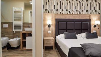 Une chambre d’hôtel avec une tête de lit en bois, un grand lit avec des oreillers, une table de chevet, une commode, une salle de bain avec des toilettes, un lavabo et un miroir.