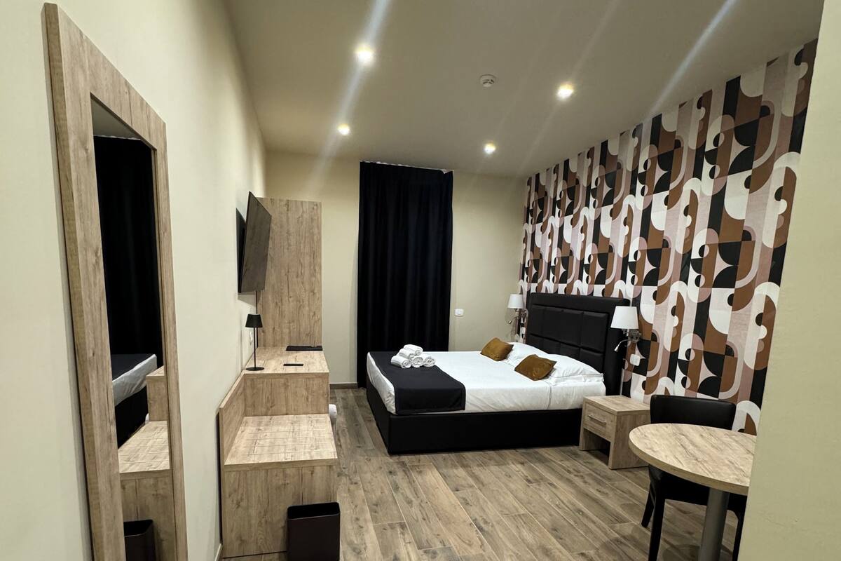 A modern hotel room with a wooden floor, a bed with black bedding, a desk with a chair, a small table, and a large mirror.