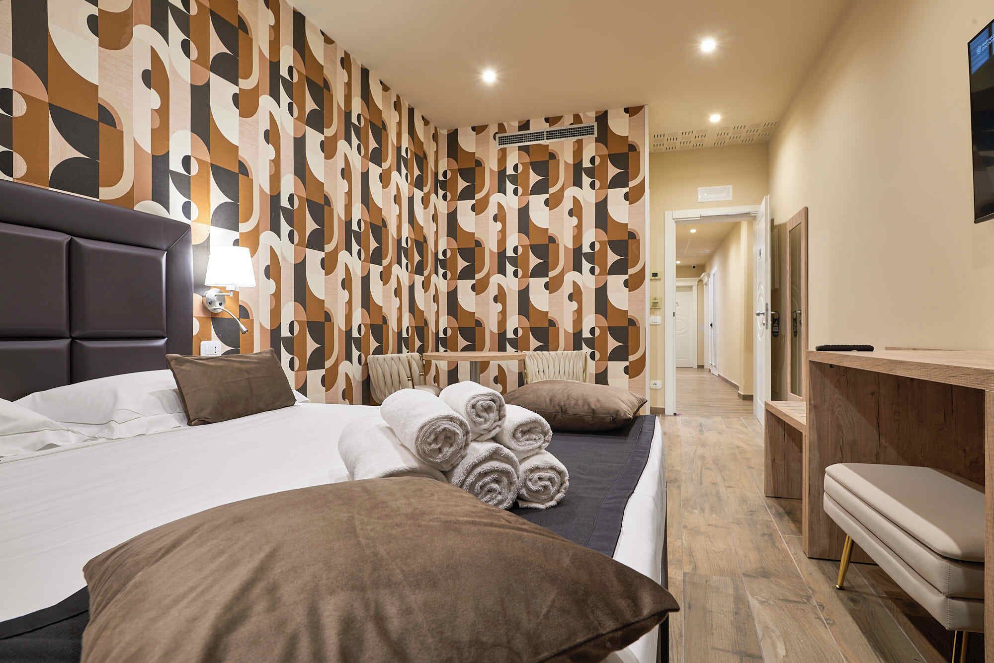 A hotel room with a large bed, a bedside table, a desk, and a chair. There is a patterned wallpaper on the wall and a door leading to another room.