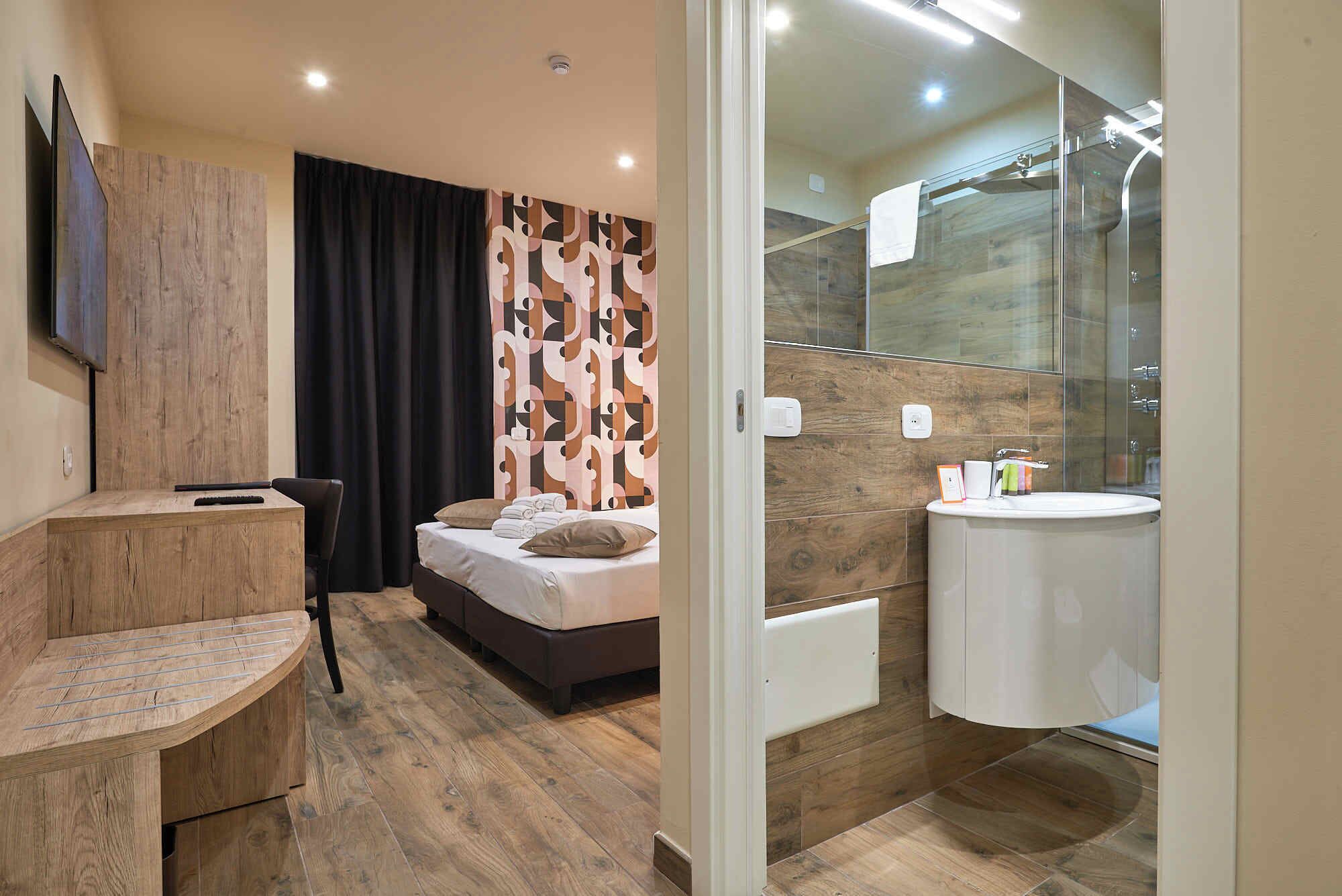Una habitación de hotel moderna con un escritorio de madera, una cama con cabecera estampada, una zona de ducha y un lavabo redondo.