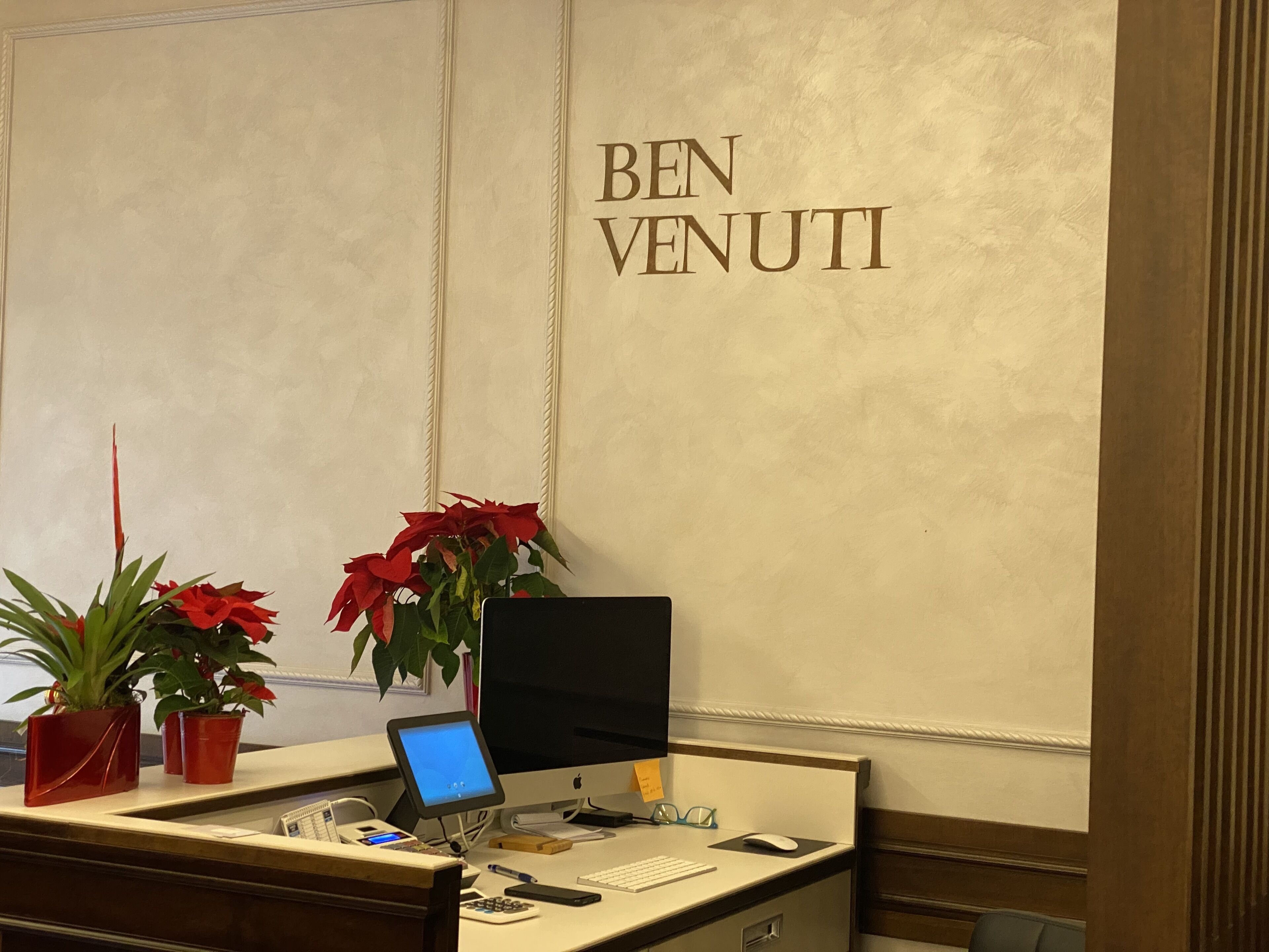 A desk with a computer, a keyboard, a mouse, a calculator, and a smartphone. There are two potted plants with red flowers. The wall has the text "BEN VENUTI" in gold letters.