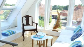 Una habitación acogedora con suelo de madera, un sofá blanco con cojines azules, una silla de madera y una pequeña mesa con un libro.
