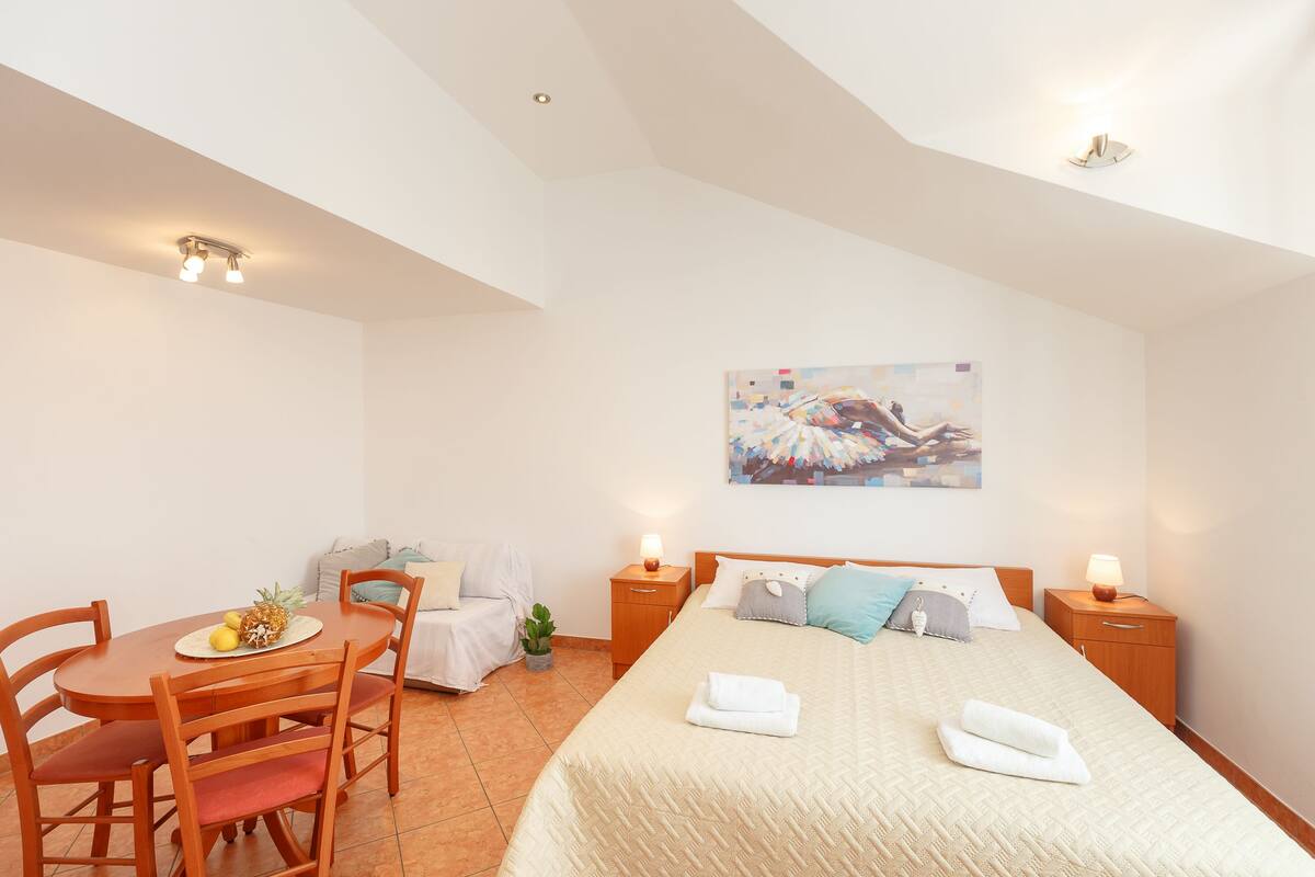 A bedroom with a bed, bedside tables, a desk with chairs, a small table with fruits, and a painting on the wall.