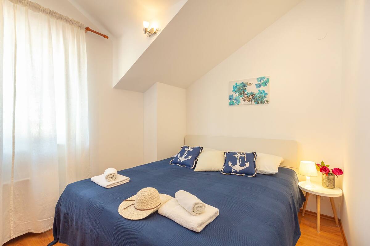 A bedroom with a bed, pillows, a nightstand, a lamp, and a painting on the wall.
