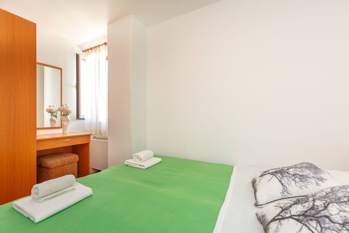 A hotel room with a green bedspread, a wooden wardrobe, a mirror, and a small table with a vase of flowers.