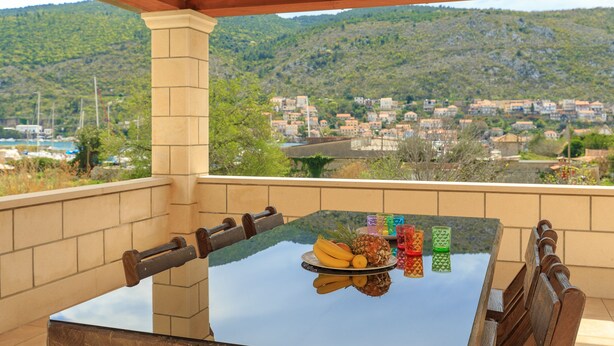 A table with a glass top, a plate of fruit, and a view of a town with houses and hills in the background. - Apartments Mondo (Dubrovnik)