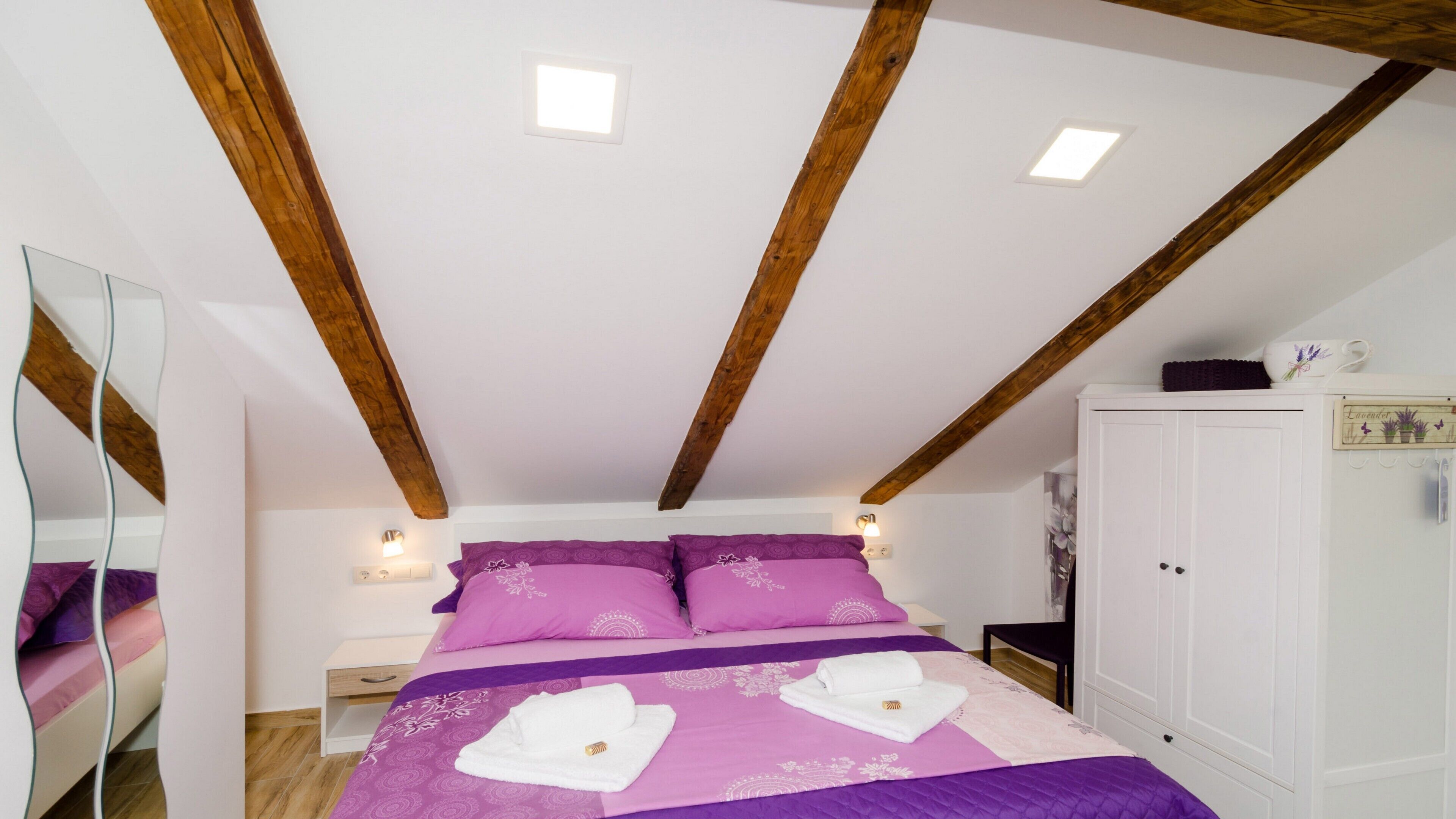 Een slaapkamer met een houten plafond, een bed met paars beddengoed, een witte garderobe en een spiegel.