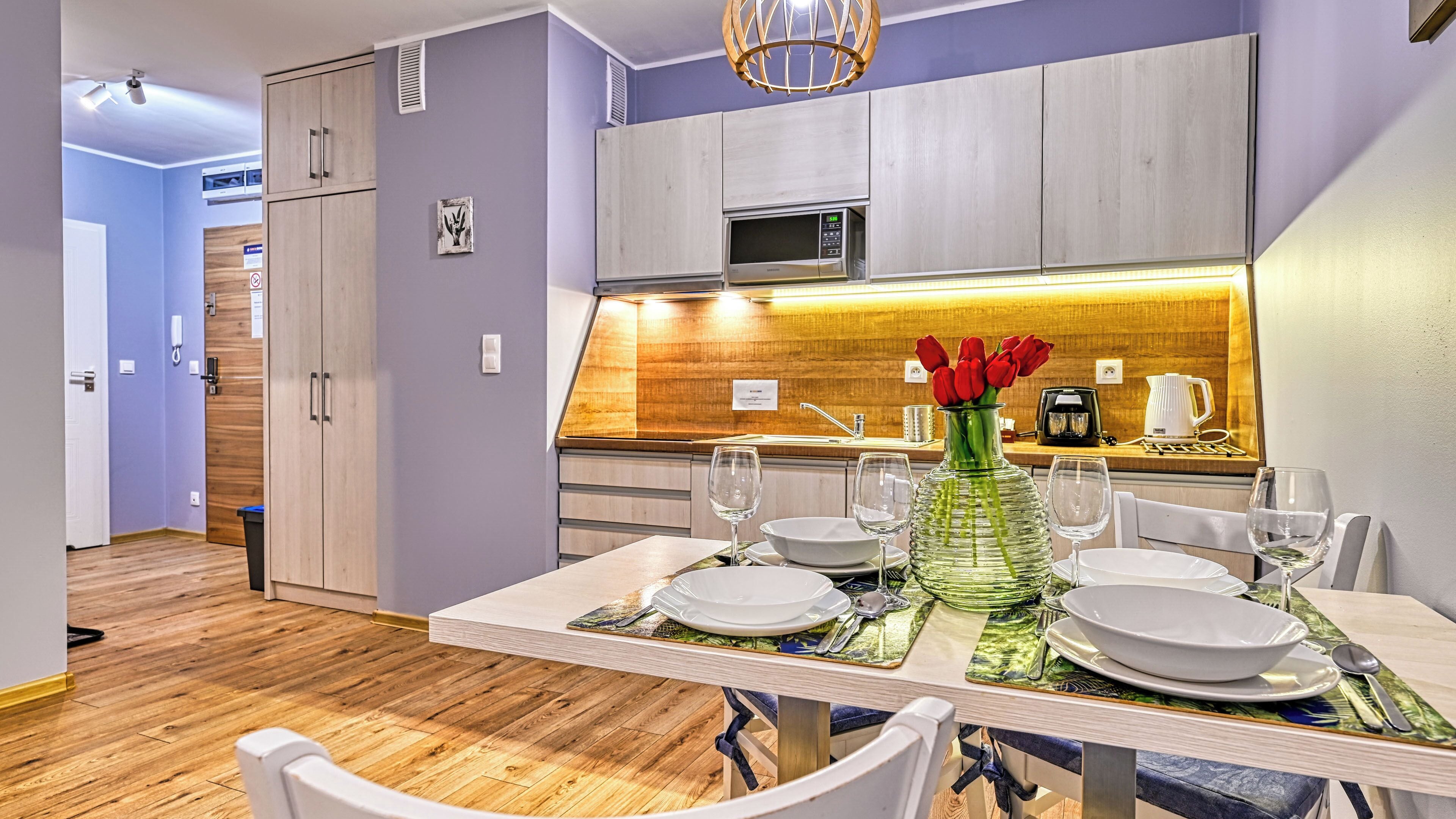 A modern kitchen with wooden cabinets, a microwave, and a sink. There is a dining table set with plates, glasses, and a vase with red flowers.