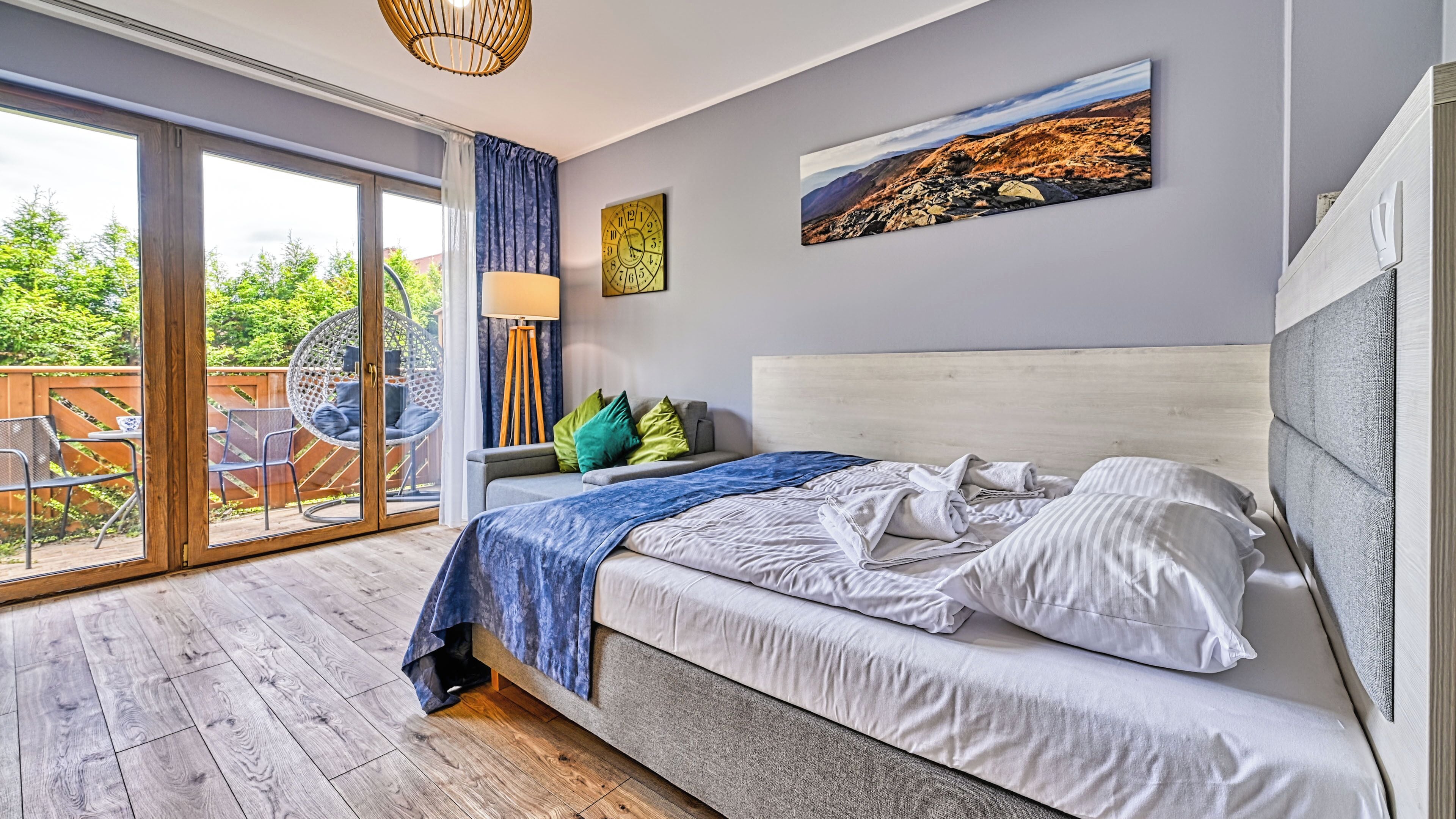 A bedroom with a bed, a view of the outdoors, a clock, and a painting on the wall.