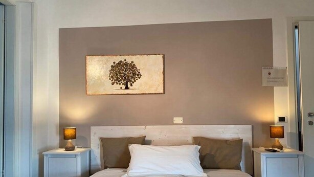 A bedroom with a bed, two bedside tables with lamps, a wall-mounted artwork of a tree, and a door on the right. - Dimora di Bosco Room & Breakfast (Rubano)