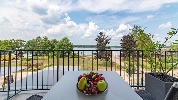 Un balcon avec une table dressée, ornée de fruits, donnant sur un lac et un parc.