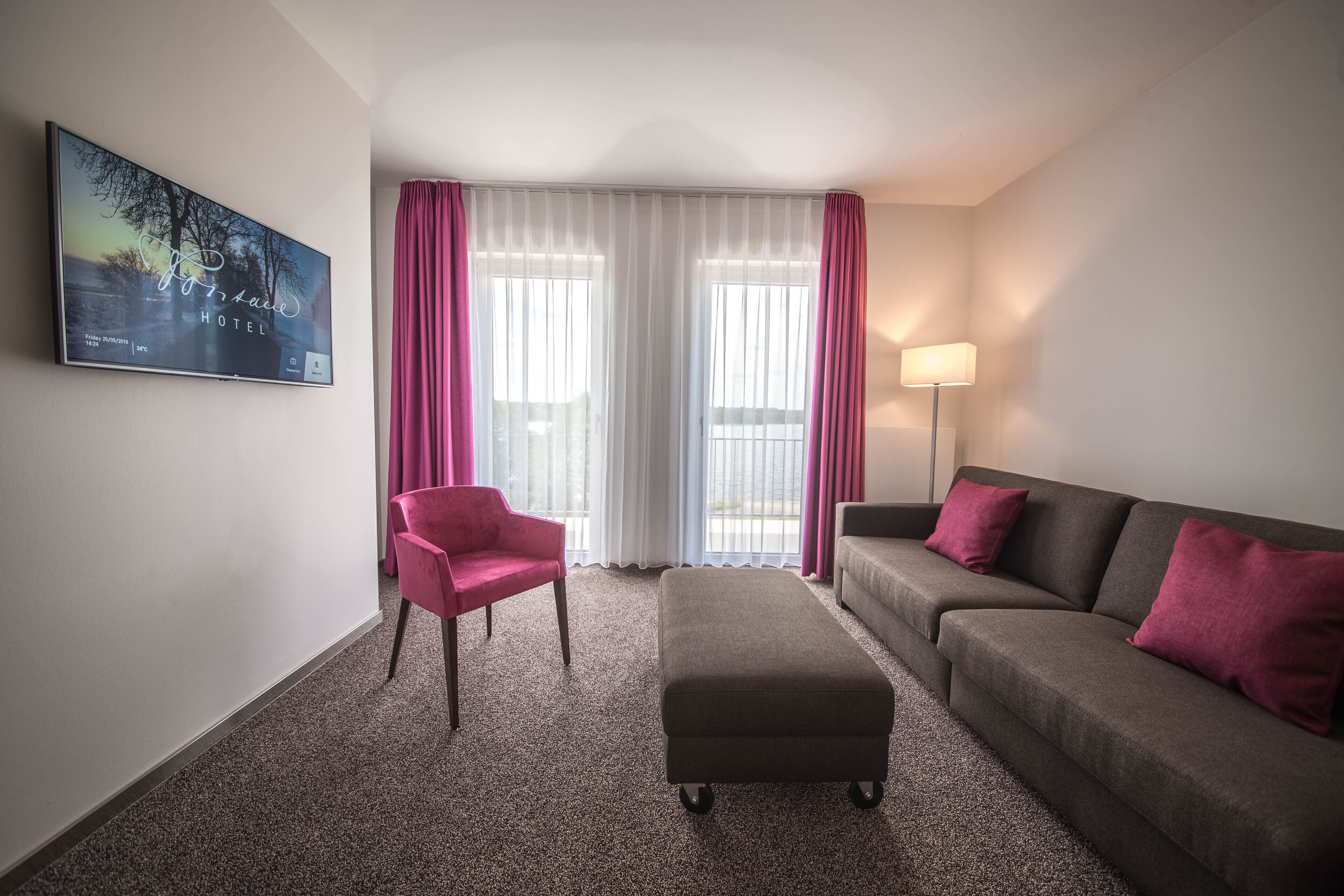 A hotel room with a large window, a flat-screen TV, a pink chair, a grey sofa, and a small ottoman.