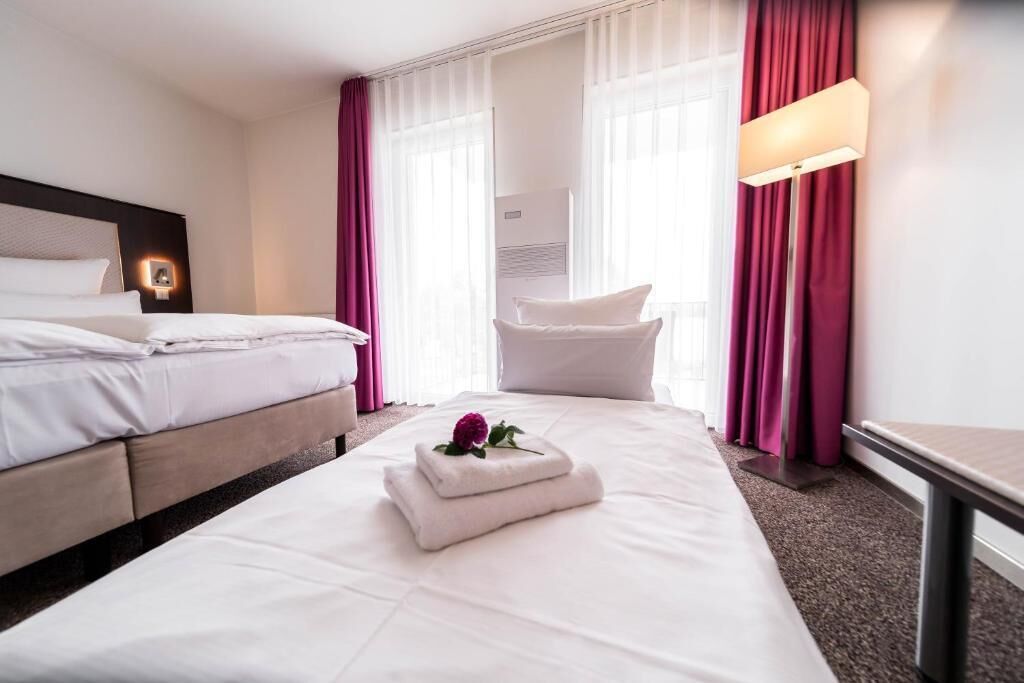 Una habitación de hotel con cama, mesita de noche, lámpara y ventana con cortinas.
