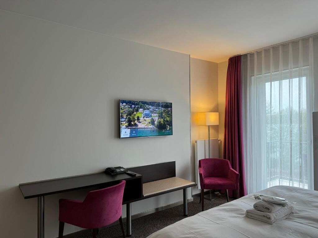 Habitación de hotel con cama, escritorio con silla, televisor montado en la pared y ventana con cortinas.
