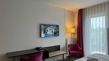 Habitación de hotel con cama, escritorio con silla, televisor montado en la pared y ventana con cortinas.