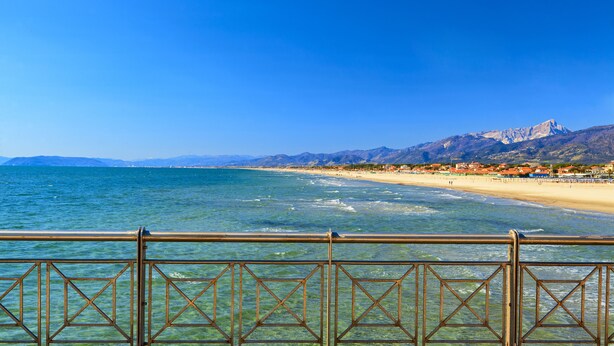 Blick auf die Küste mit einer Holzterrasse und Metallgeländer, das auf einen Strand und Berge blickt.