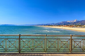 A coastal view with a wooden deck and metal railing, overlooking a beach and mountains.