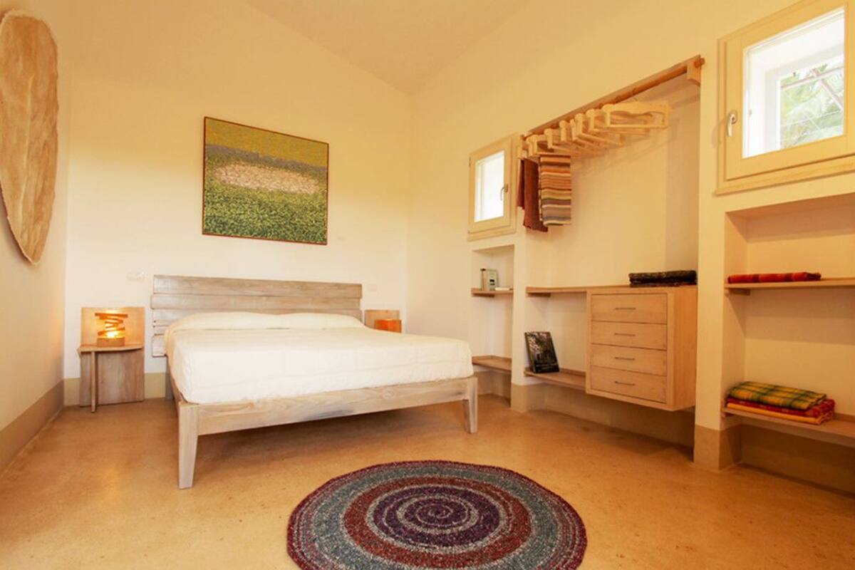 Une chambre avec un lit en bois, une table de chevet avec une lampe, une étagère murale avec des serviettes et un tableau au-dessus du lit.