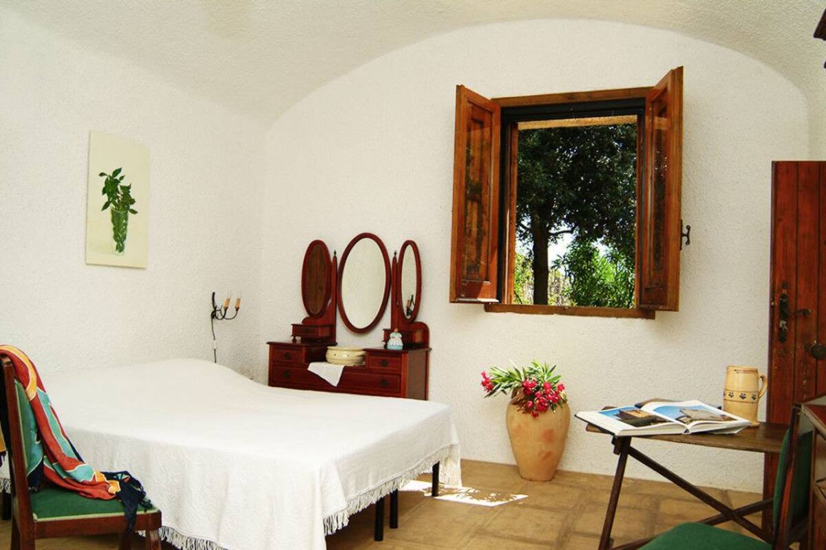 Une chambre à coucher avec un lit, une commode, un miroir, une fenêtre donnant sur des arbres, une chaise, une table avec un livre et un vase de fleurs.