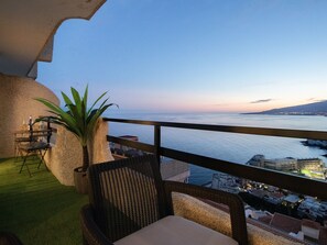 Un balcon avec un fauteuil en osier et une plante, donnant sur un paysage urbain côtier au crépuscule.