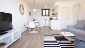 Ein modernes Wohnzimmer mit einem Flachbildfernseher, einem grauen Sofa, einem Streifenmuster-Teppich und einem Essbereich mit weißen Stühlen und einem Tisch.