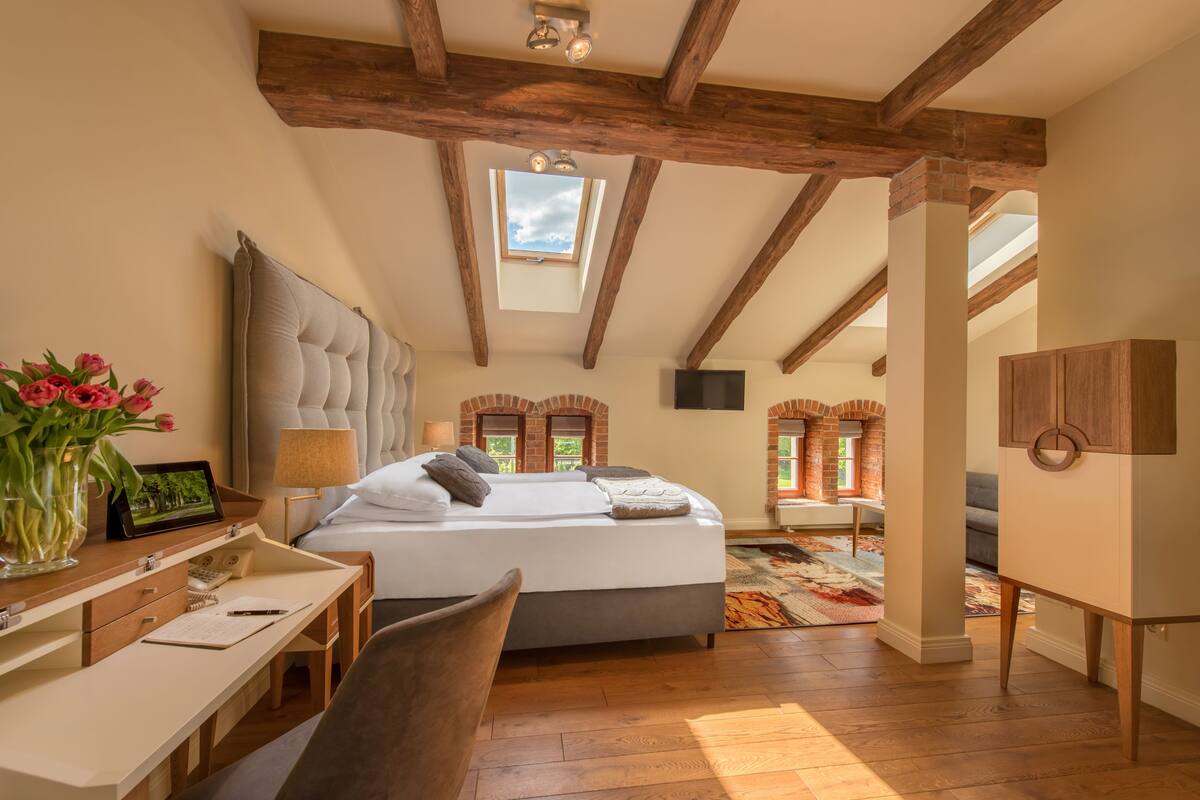 A bedroom with a large bed, wooden beams, a skylight, a desk with a lamp, and a view of a brick wall and windows.
