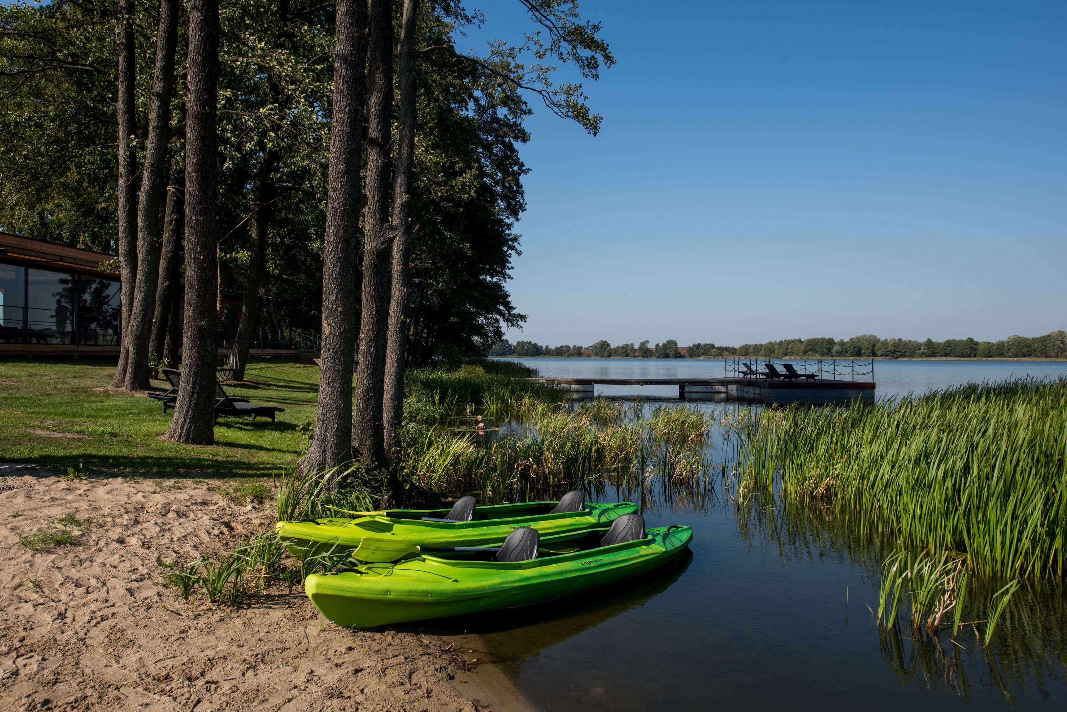 A lakeside area with green kayaks, a dock, and a building in the background.