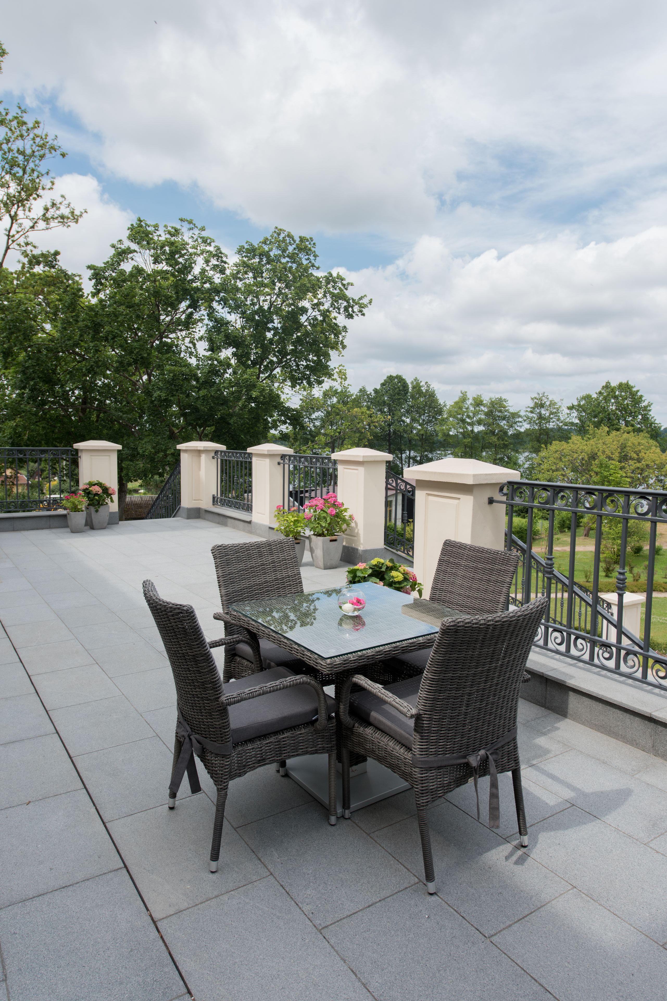 A terrace with a glass table and wicker chairs, surrounded by potted plants and a metal railing.