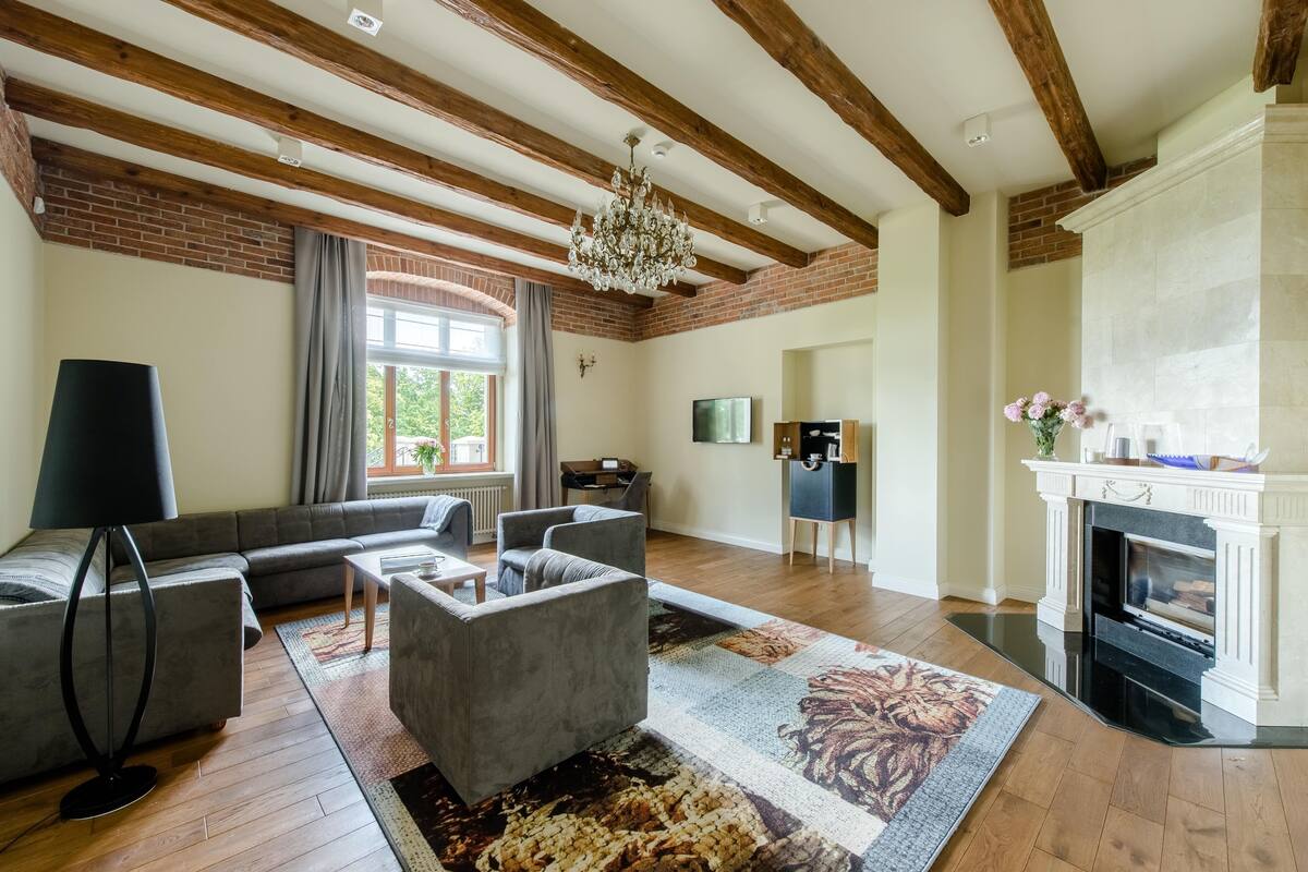 A spacious living room with a fireplace, a large window with curtains, and a modern chandelier.