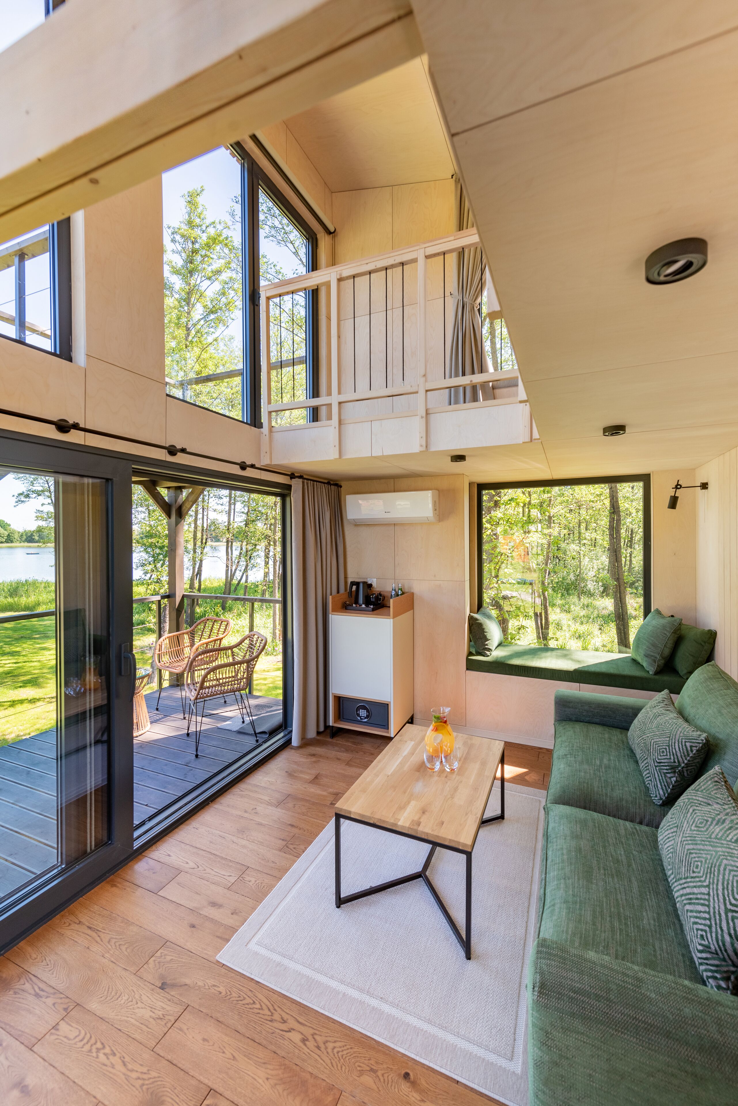 A spacious living area with a wooden floor, a green sofa, a wooden coffee table, wicker chairs, and a view of a lake through large windows.