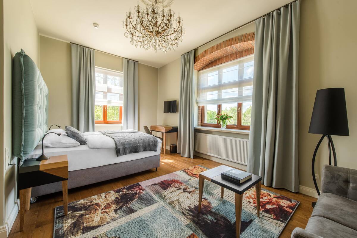 A modern bedroom with a bed, a desk, a chair, a lamp, and a view of the outdoors through large windows.