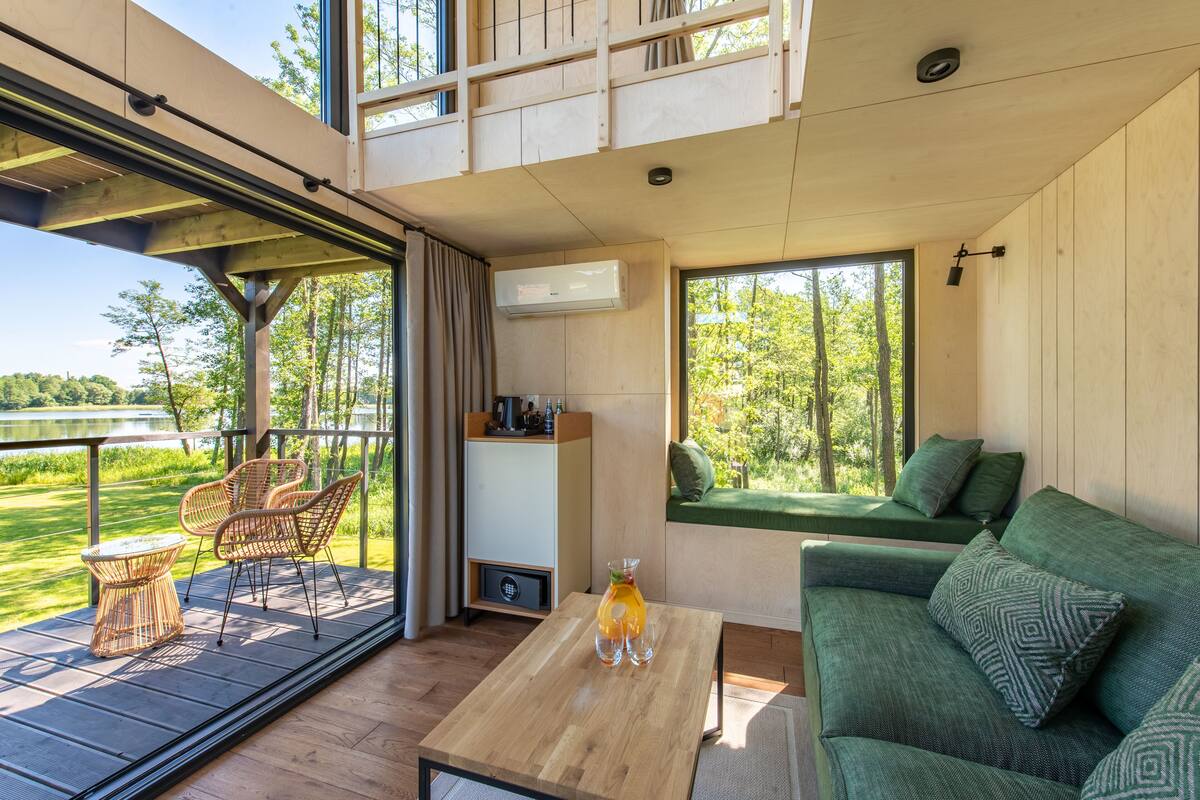 A modern room with a wooden floor, a green sofa, a wooden coffee table, wicker chairs, and a large window overlooking a garden.