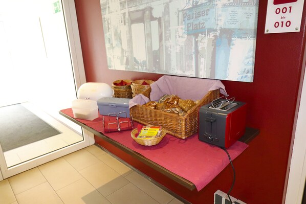 Un área de recepción con una pared roja, una pantalla digital que muestra una vista de una calle parisina, una mesa roja con cestas, un tostador y un cesto de pan.