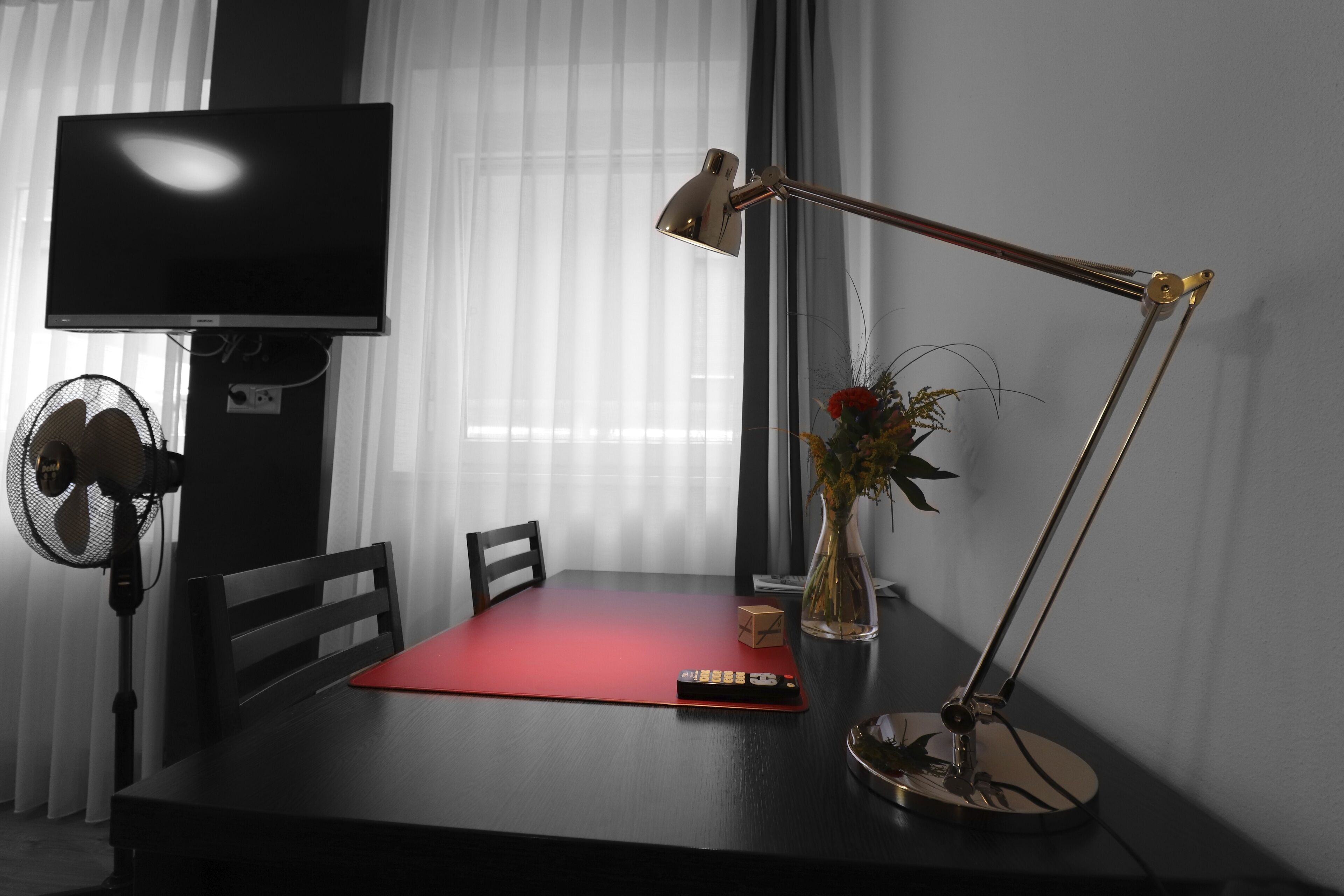 Una habitación con una televisión en la pared, un escritorio con lámpara, una silla, una mesita y un ventilador.