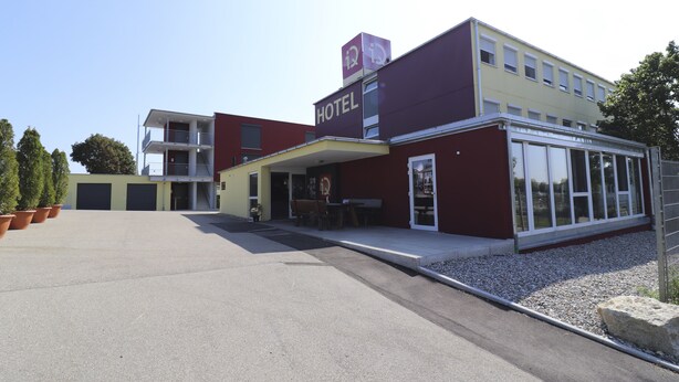 A modern hotel with a flat roof, a covered entrance, and a visible outdoor seating area. - IQ Hotel (Langenau)