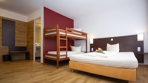 A room with a bunk bed, a single bed, wooden flooring, a red accent wall, a wooden headboard, and a small table. - IQ Hotel (Langenau)