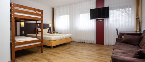 A room with a bunk bed, a single bed, a brown sofa, a TV mounted on the wall, and a window with sheer curtains.