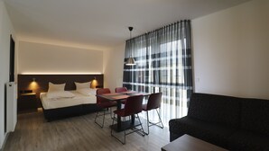 A modern hotel room with a bed, a dining table with chairs, a sofa, and a TV. - IQ Hotel (Langenau)
