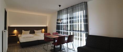 A modern hotel room with a bed, a dining table with chairs, a sofa, and a TV.