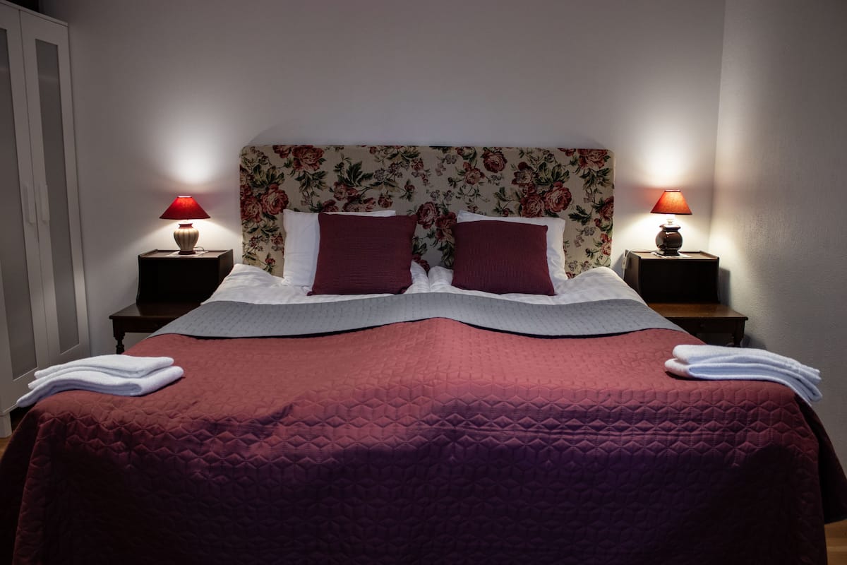 A neatly made bed with a floral headboard, two bedside lamps, and folded towels on the bed.
