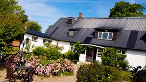 A two-story house with a dark roof, white walls, and a small balcony. There is a garden with pink flowers and a table with chairs in the foreground. - Stora Herrestad Bed & Breakfast (Ystad)