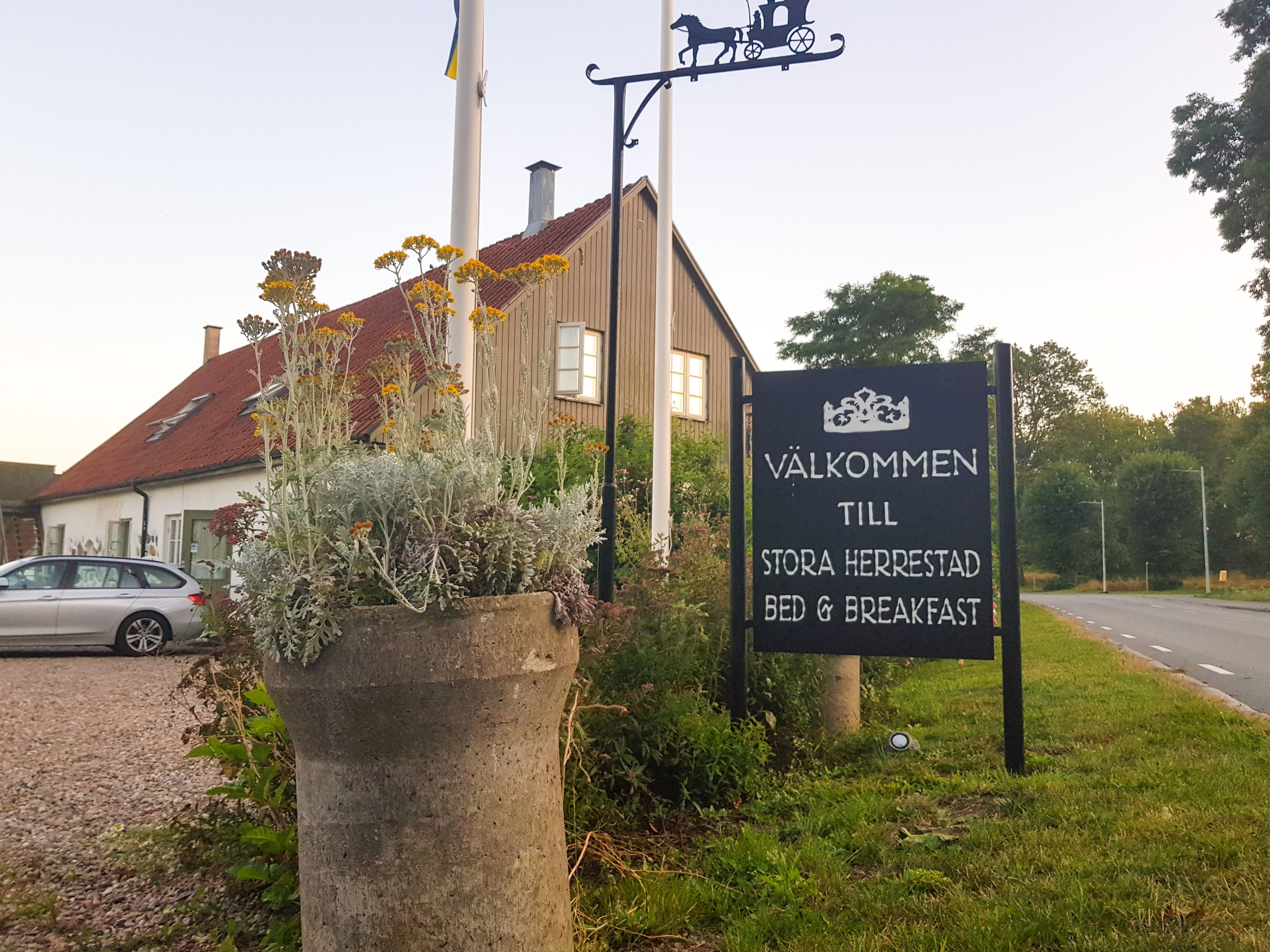 Señal de un bed and breakfast llamado Valkommen, situado cerca de una carretera con un coche estacionado.