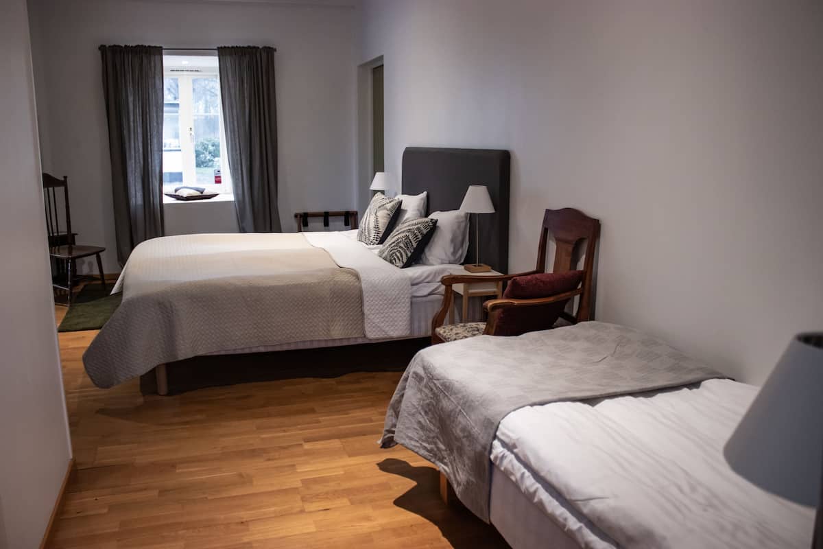 A hotel room with two beds, a wooden floor, a window with curtains, and a chair.