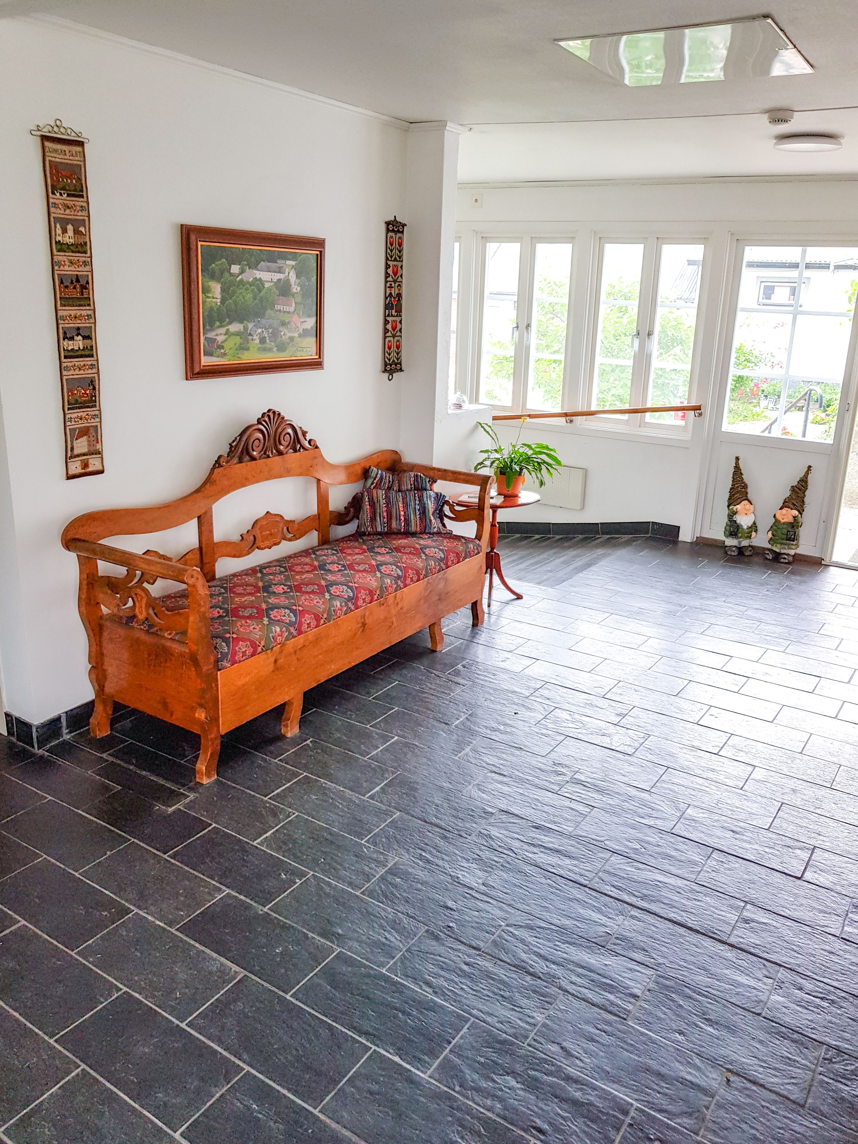 A wooden sofa with a patterned cushion in a room with dark tiled flooring, a large window, and a plant on a small table.